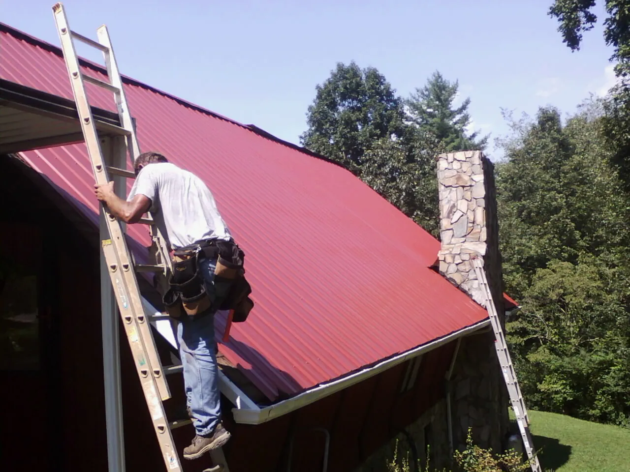 Roofer on a ladder beside a red metal roof and stone chimney — Soffit & Fascia Repair by Roofing Brothers in Nolensville